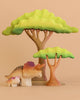 The Bumbu Handmade Wooden Prehistoric Set features an adult and baby dinosaur with two stylized acacia trees, crafted from wood to inspire imaginative and educational play on a beige background.