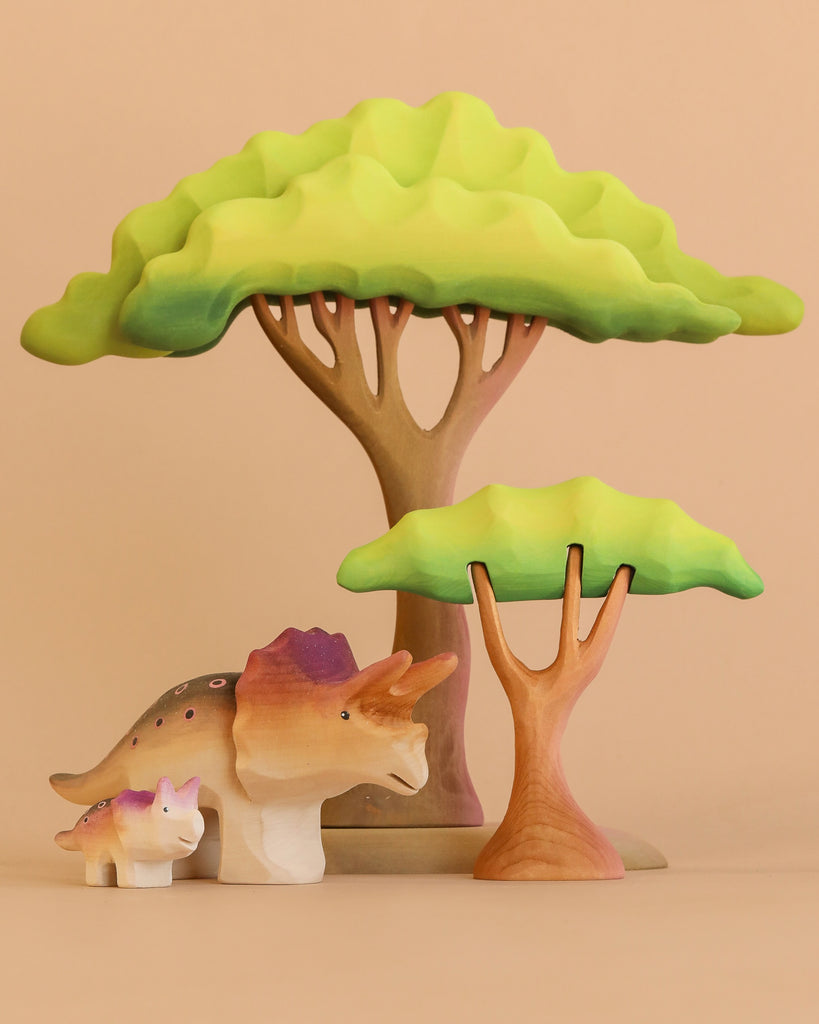 The Bumbu Handmade Wooden Prehistoric Set features an adult and baby dinosaur with two stylized acacia trees, crafted from wood to inspire imaginative and educational play on a beige background.