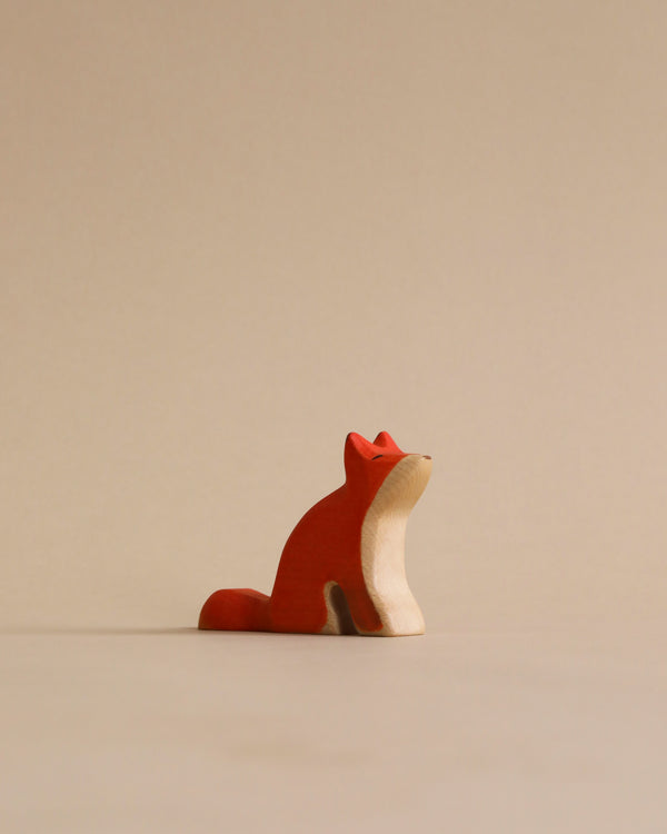 A sustainable Handmade Holzwald Sitting Fox figurine sits elegantly against a plain light beige background, its head tilted upwards as if gazing at something in the distance.