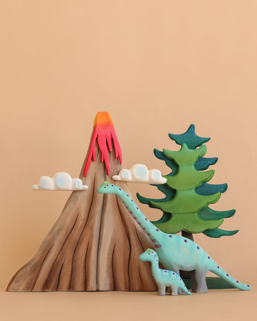 The Bumbu Handmade Wooden Prehistoric Set includes a toy volcano with red lava, white clouds, two pine trees, and blue and green dinosaurs—all painted with non-toxic paint for safe, imaginative play against a beige background.