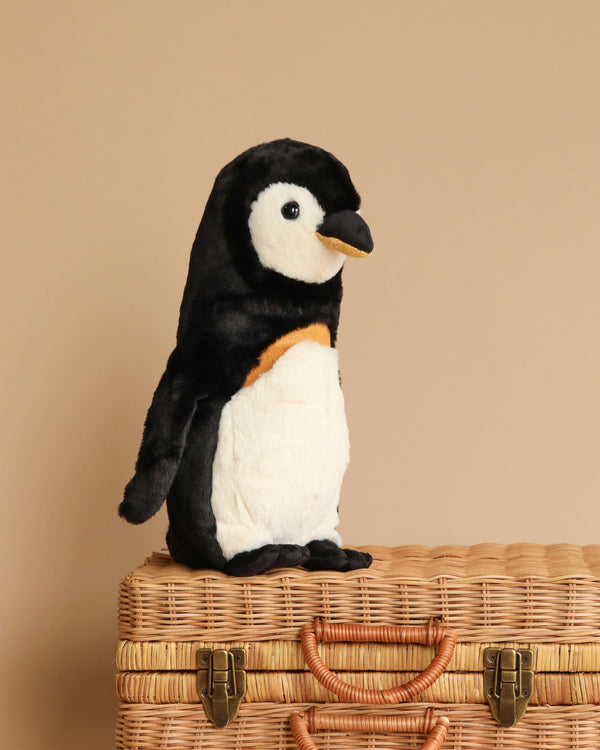 A Nala Penguin Stuffed Plush Animal, 13 inches tall, is sitting on a closed wicker basket against a plain beige background. The penguin's upright body showcases its soft texture and realistic black and white fur.