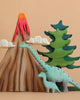 Two figures from the Bumbu Handmade Wooden Prehistoric Set stand before a volcano with red lava, fluffy clouds, and a green pine tree—perfect for imaginative play against a beige backdrop.