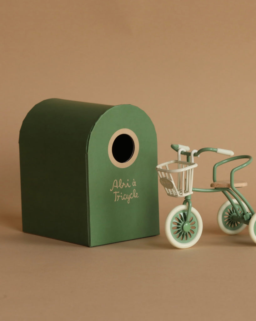 The Maileg Tricycle With Basket - Green stands beside a small green box labeled "Abri à Tricycle," both set against a plain beige background. The tricycle features a front basket and green and white detailing.