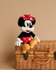 An image of the Steiff, Disney's Minnie Mouse Stuffed Plush Toy, 12 Inches sitting on a wicker basket. The plush toy, part of the Disney Originals Soft Cuddly Friends Collection, is wearing a red dress with white polka dots, a matching bow, and yellow shoes. The background is a plain beige color.