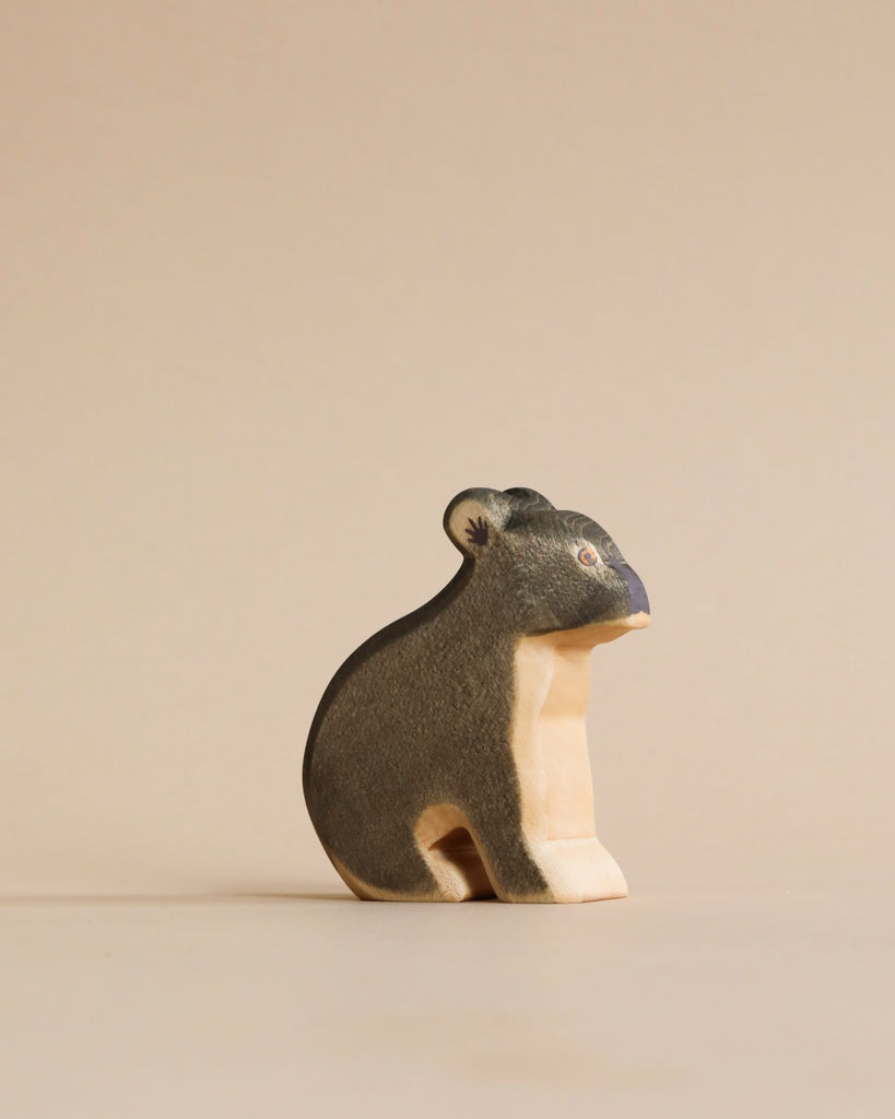 A Handmade Holzwald Koala, painted in dark and light tones, sits against a plain, light beige background. The bear has detailed eyes and a calm posture. This piece represents sustainable toys crafted