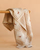 The Organic Cotton Baby Blanket - Pony, beige with a checkered pattern and small embroidered red horses, is draped over a wooden surface against a light peach background.