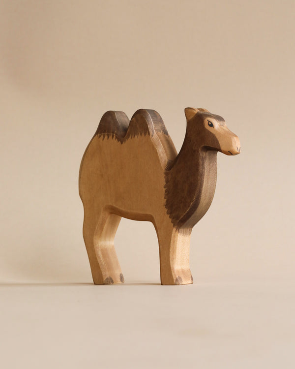 A Handmade Holzwald Camel figurine with two humps stands against a plain, light beige background. The camel, representing high-quality toys, is carved with visible wood grain and variations in its brown shading.
