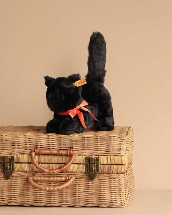 The Spooky Black Cat Plush Toy Halloween, featuring a red ribbon around its neck and tagged on its ear, showcases its cuddly softness as it sits atop a closed wicker basket with leather straps against a beige background. This small plush black cat makes the perfect addition to your Halloween decor.