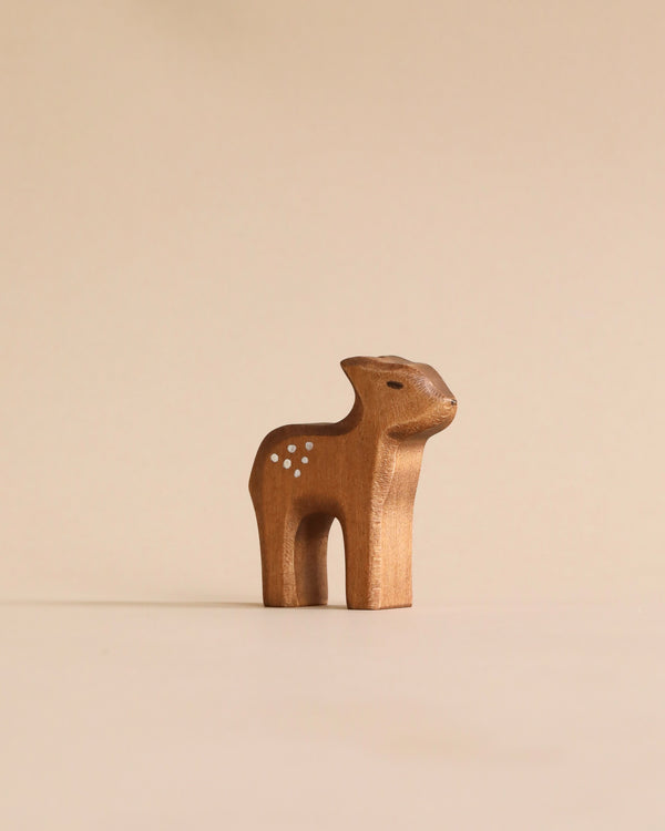 A Handmade Holzwald Standing Fawn figurine with dotted spots and pricked ears stands against a plain, light beige background, representing a delightful example of sustainable toys.