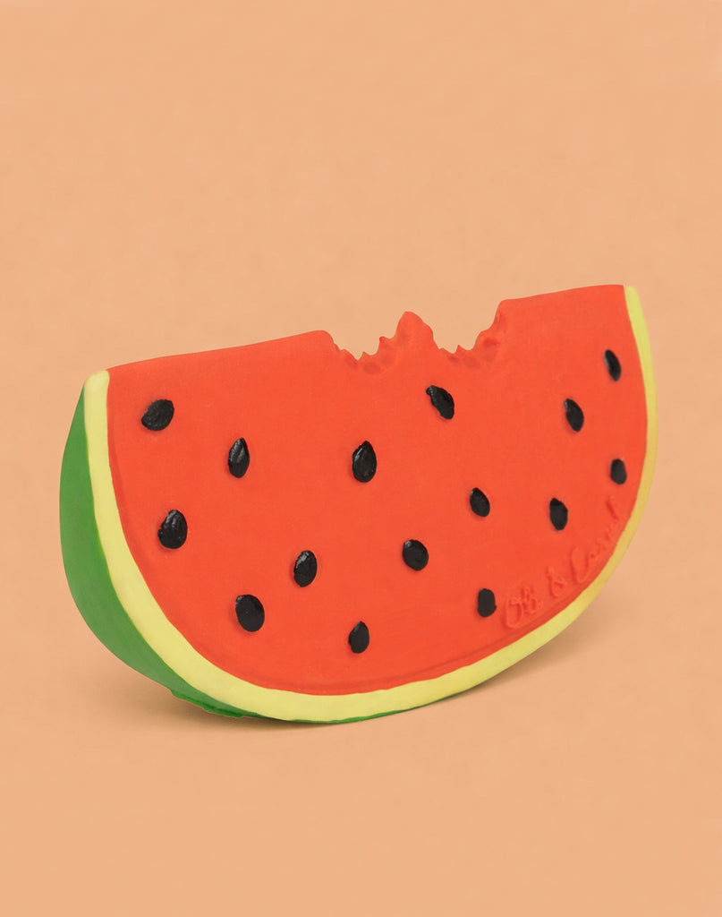 Wally the Watermelon Teether is a natural rubber baby toy shaped like a bitten watermelon slice, with a green rind, red flesh, and black seeds—ideal for sensory play—shown on a plain peach background.