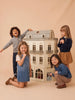 Four young children smile and pose around the House of Paris Luxury Dollhouse – A Tale of Parisian Magic, set against a beige background. Two kneel in front while two stand behind, all looking happy and engaged with the fully furnished, deluxe Parisian dollhouse.