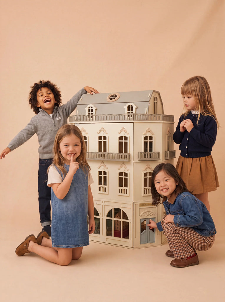 Four young children smile and pose around the House of Paris Luxury Dollhouse – A Tale of Parisian Magic, set against a beige background. Two kneel in front while two stand behind, all looking happy and engaged with the fully furnished, deluxe Parisian dollhouse.