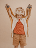 Smiling child wearing the Costume - Lion faux fur hood with raised paws, peach shirt, cream overshirt, and tan shorts, standing against a plain beige background.