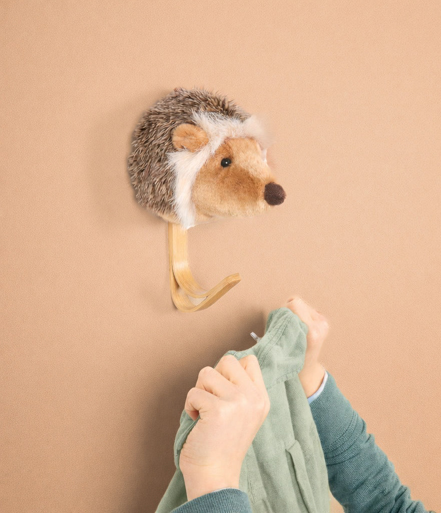 A person hangs a green shirt on the Coat Hanger - Hedgehog, a plush animal head-shaped hanger, mounted against a beige background.