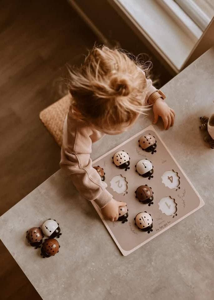 A young child plays with the My Little Lady Bug Wooden Puzzle on a table near a window, reaching to place a ladybug-shaped piece into the board.