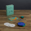 Board Game: Catch Phrase Vintage Bookshelf Edition displayed on a wooden surface, includes a teal box, scorecard, colorful markers, circular word disks, and a blue and black electronic game unit.