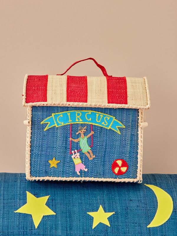 Introducing the Raffia Kids Bag with Circus Theme—a small, handmade box crafted from raffia. It features a cheerful red-and-white striped roof, a blue front, and embroidered animals swinging in playful motion. Resting on a blue cushion adorned with yellow stars and a crescent moon, this sustainable piece doubles as both an adorable kids bag and whimsical decor for any room.