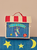 Introducing the Raffia Kids Bag with Circus Theme—a small, handmade box crafted from raffia. It features a cheerful red-and-white striped roof, a blue front, and embroidered animals swinging in playful motion. Resting on a blue cushion adorned with yellow stars and a crescent moon, this sustainable piece doubles as both an adorable kids bag and whimsical decor for any room.