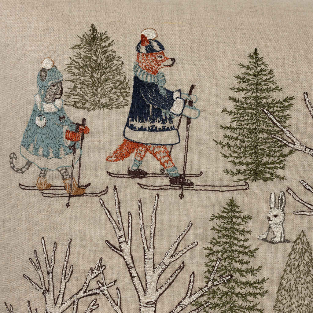 The Coral & Tusk Winter Fun Pillow features embroidered woodland animals—a cat and a fox—in winter outfits skiing through a snowy forest, with evergreen trees, a white rabbit, and festive details, perfect for holiday decor.