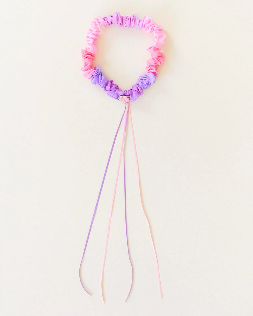 A Sarah's Silk Blossom Garland Headband is pink and purple with long dangling ribbons, reminiscent of festive garlands. The headband is ruched and the ribbons extend from the bottom, adding a decorative touch. Perfect for dress-up play, it is set against a plain, light background.