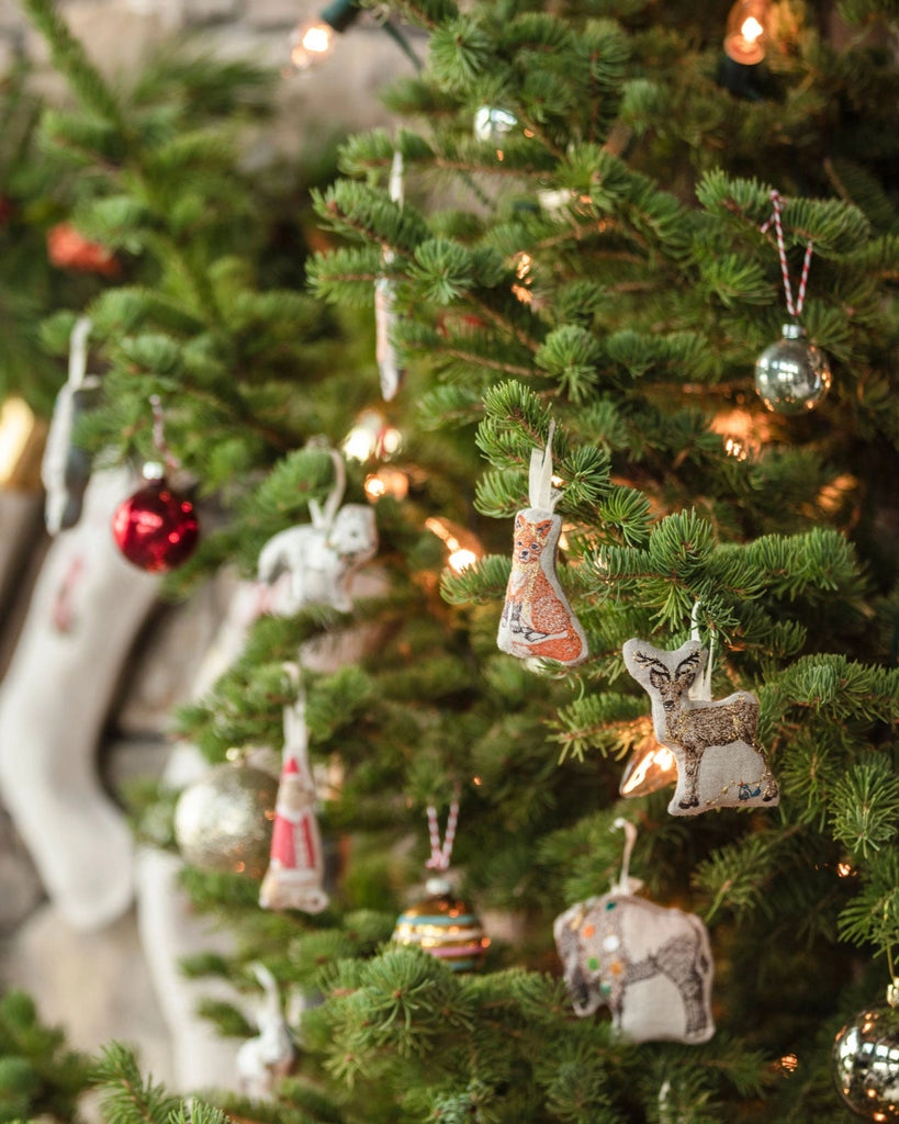 A Christmas tree adorned with animal-shaped ornaments, including the Coral & Tusk Fox Tree Trimmer Ornament, owl, and elephant, along with round baubles and glowing lights—a festive holiday gift with a cozy, softly blurred background.