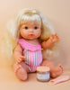 A blonde baby doll wearing a pink and white striped outfit sits on a beige surface, poised for her meal. Next to her, the "Tiny Tummies - Coconut Yogurt" jar from Tiny Harlow and its magic spoon are ready for use. The soft peach background creates an idyllic setting as the doll enjoys her delightful dining experience.