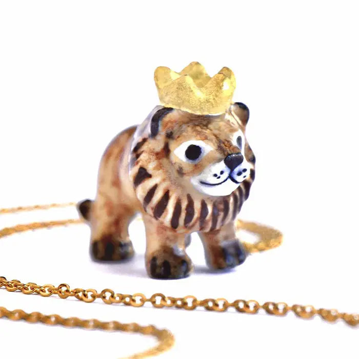 A small figurine of a lion wearing a gold crown is depicted. Crafted from high-quality porcelain, the Lion King Necklace boasts detailed fur patterns and a playful, cartoonish style with a cheerful expression. It stands on a white surface, surrounded by a delicate gold chain, showcasing hand-painted details.