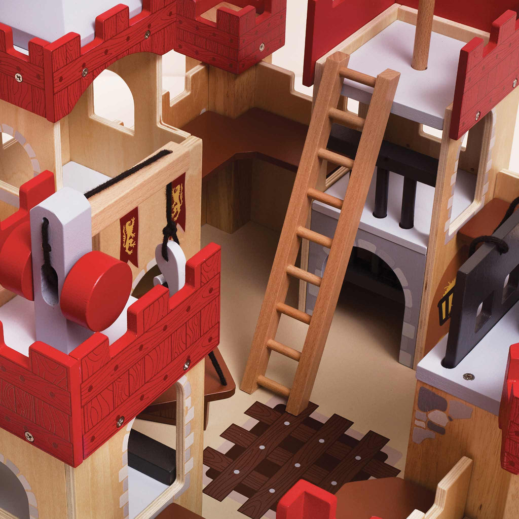 A close-up of the Lionheart Wooden Castle, beautifully crafted from sustainable wood and featuring vibrant red battlements, a working drawbridge, a ladder, banners adorned with lion emblems, as well as detailed windows and a dungeon gate. This colorful structure is designed to spark imaginative play.