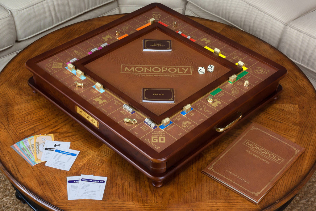 The Monopoly Luxury Edition, a leather-bound board with gold accents and die-cast metal pieces, sits on a wooden table alongside cards, dice, Monopoly money, and a matching leather-bound rulebook.