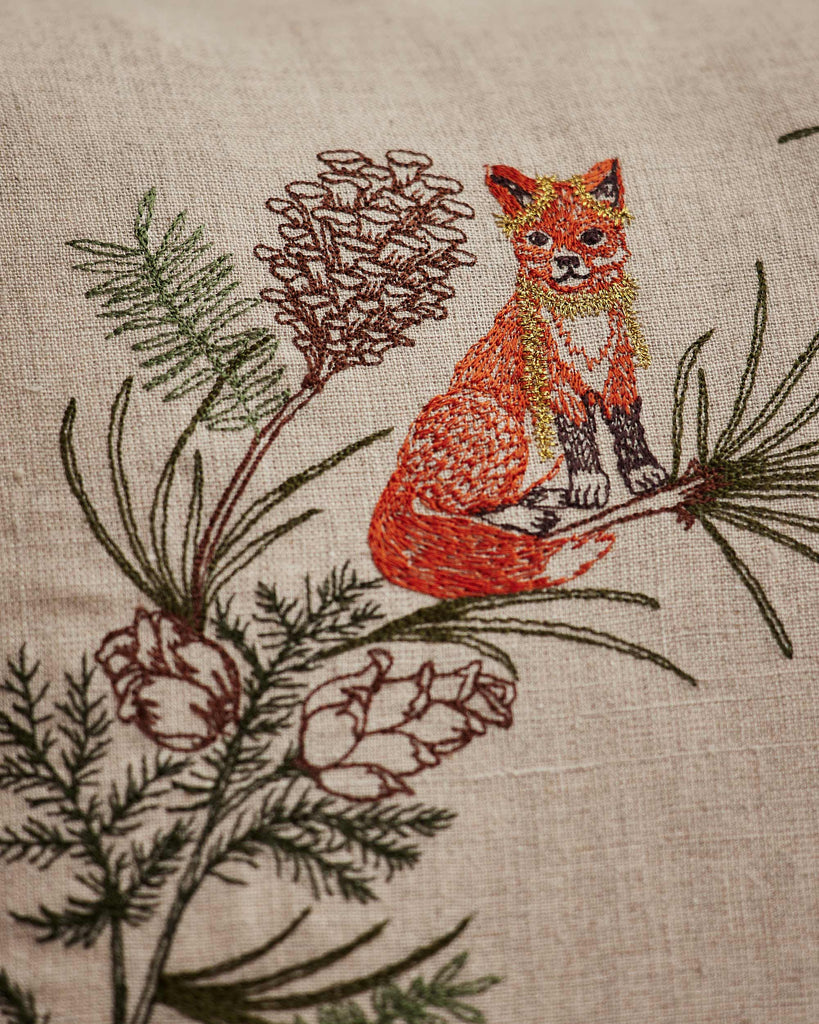 The Coral & Tusk Tree Trimmers Wreath Pillow showcases an embroidered orange fox on a branch with pinecones, green pine needles, and twigs—ideal as a cozy animal pillow for festive Christmas decor.