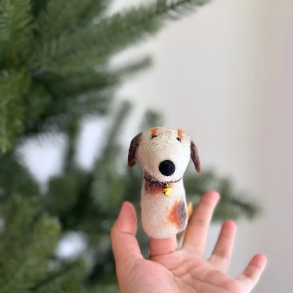 A hand holds up a Calico Dog Finger Puppet, a small needle-felted stuffed dog ornament with a white body and brown spots, in front of a blurred Christmas tree. The dog features closed eyes, floppy ears, and wears a tiny yellow bell collar. Lovingly handmade in Nepal, it's perfect as a unique finger puppet.