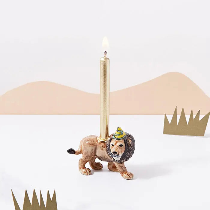 A unique Lion Cake Topper shaped like a lion, crafted from fine porcelain, featuring a lit slender taper candle on its back, positioned against a minimalist background with abstract brown mountains and green cutouts.