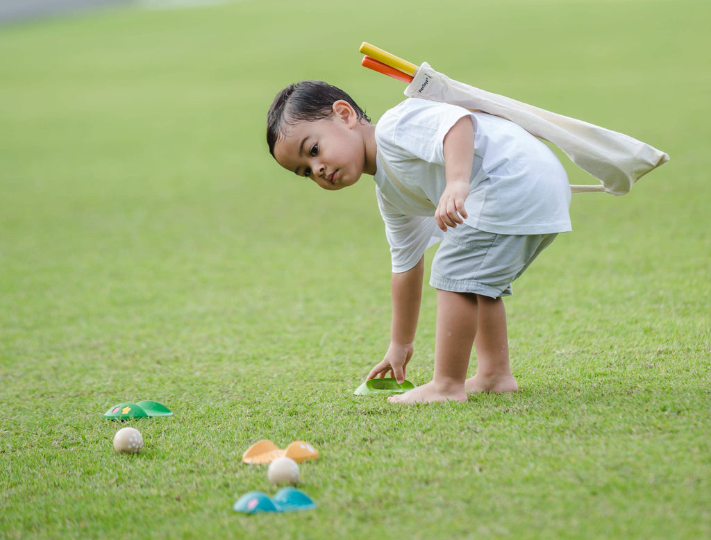 A young child bends down on the grass to pick up a green marker while carrying the Mini Golf Set over their shoulder; balls and cone markers are scattered across the lawn for this fun backyard golf game.