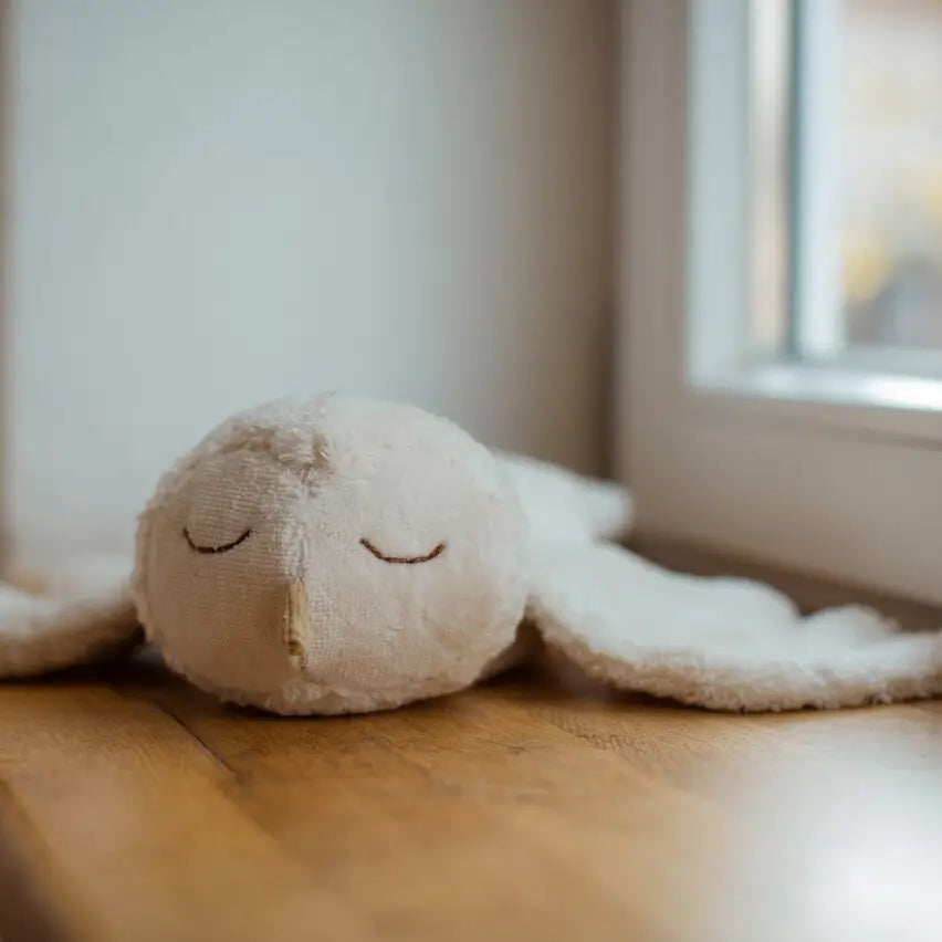 The Senger Naturwelt Cuddly Animal - Owl, a soft white plush toy with stitched eyes and beak, rests on a wooden floor by the window.
