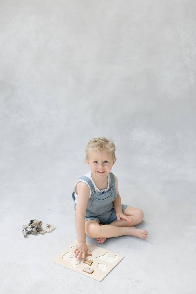 A young child with blonde hair sits on the floor, smiling, while playing with the Farm Animals Wooden Puzzle: A Tabitha Paige Puzzle, which features friendly animals. Several puzzle pieces are scattered nearby, making this engaging educational toy both fun and beneficial for toddlers. The background is light and neutral.