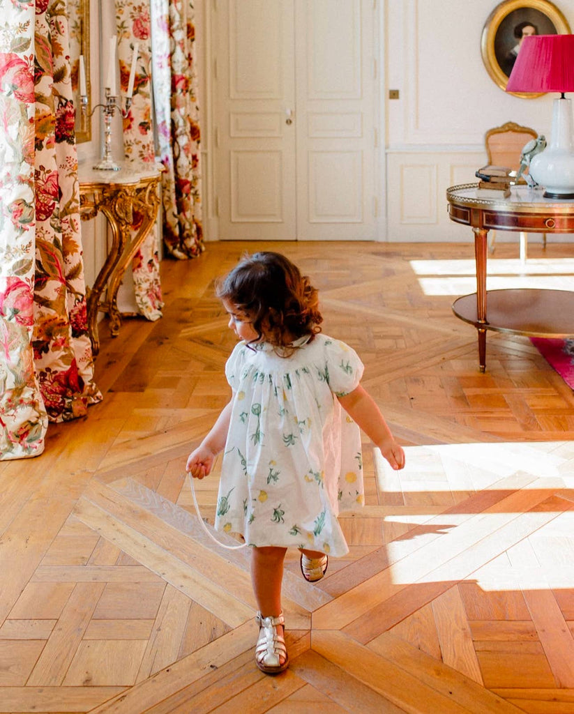 A young girl in a white floral dress walks barefoot, gently pulling her Pull-Along Walter with its wooden wheels across a patterned wooden floor in a bright, elegant room adorned with floral curtains, ornate furniture, and a round table topped with a pink lamp. This charming scene not only highlights her innocence but also encourages the development of her gross motor skills.