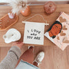 A person holds the I Love Who You Are Canvas Sign - Natural on a wooden table decorated with jewelry, Kenyan artisan decor, a vase, pink cup, and an illustrated book; they wear white sneakers.