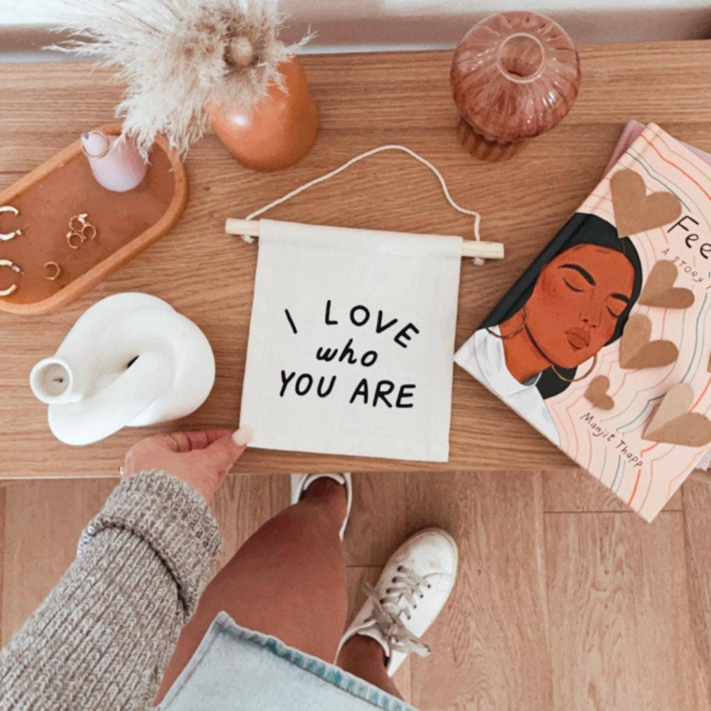 A person holds the I Love Who You Are Canvas Sign - Natural on a wooden table decorated with jewelry, Kenyan artisan decor, a vase, pink cup, and an illustrated book; they wear white sneakers.