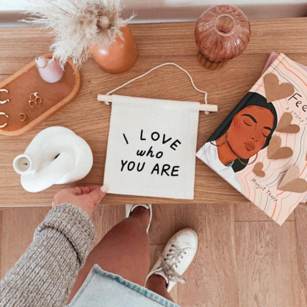 A person holds the I Love Who You Are Canvas Sign - Natural on a wooden table decorated with jewelry, Kenyan artisan decor, a vase, pink cup, and an illustrated book; they wear white sneakers.