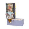 The Moulin Roty Pablo the Elephant - The Minouchkas sits in patterned clothes on a purple box, with a matching open case behind it that features vibrant floral designs and an illustrated house background.