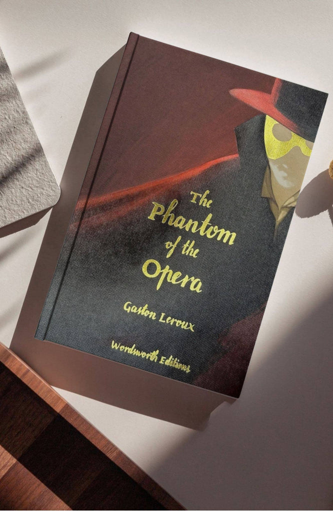 A hardcover collector’s edition of “The Phantom of the Opera” featuring Gaston Leroux’s classic horror tale, with a cover illustration depicting a masked figure in a red hat and cape, displayed on a light background with shadow patterns.