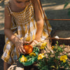 A young child in a yellow plaid dress picks a red tomato and places it in the green tray from the Kendall the Kale Plate & Spoon Set, surrounded by yellow flowers in a garden.