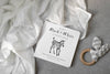 The Black and White Contrast Book for Baby, featuring a zebra on the cover, rests on a soft white cloth beside a gray striped fabric and a wooden teether, all arranged on a light gray textured surface. Designed with high-contrast images, this book supports your little one’s early visual development.