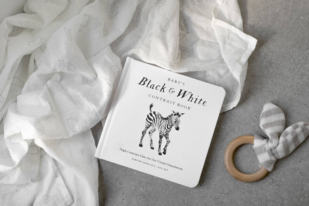 The Black and White Contrast Book for Baby, featuring a zebra on the cover, rests on a soft white cloth beside a gray striped fabric and a wooden teether, all arranged on a light gray textured surface. Designed with high-contrast images, this book supports your little one’s early visual development.