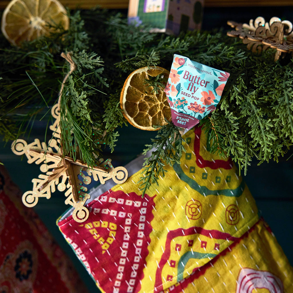 A close-up of a colorful Christmas stocking hanging on a garland with dried orange slices, a wooden snowflake ornament, and a Pollinator Seed Pop (Single)—a thoughtful gift for anyone dreaming of starting a pollinator garden.