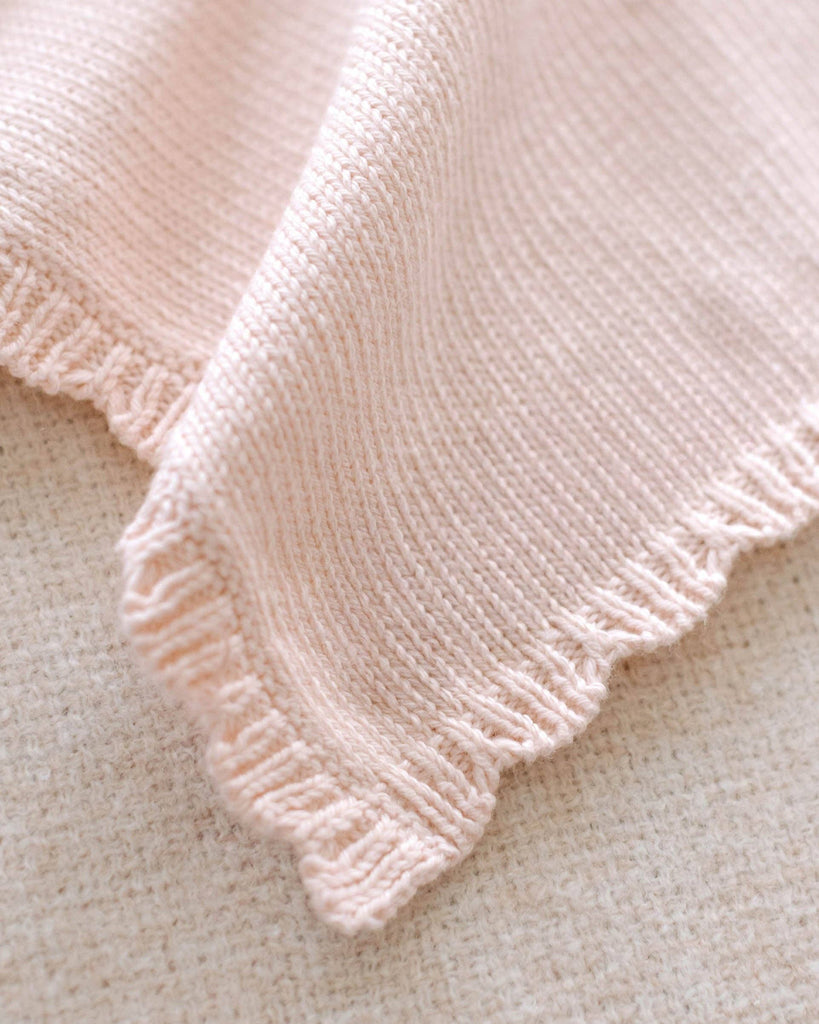 A close-up of the Cuddle + Kind Organic Cotton Baby Bear Lovey (Blush), featuring soft, hand-knit light pink fabric and scalloped edges, draped over a textured beige surface.