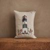 The Coral & Tusk Lighthouse Friends Pocket Pillow—beige linen with embroidered lighthouse, animals, and plants—rests on a wooden surface against a textured brown wall.