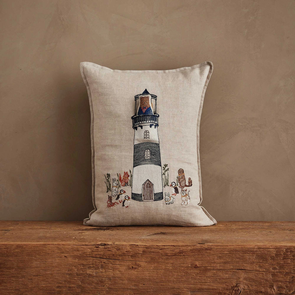 The Coral & Tusk Lighthouse Friends Pocket Pillow—beige linen with embroidered lighthouse, animals, and plants—rests on a wooden surface against a textured brown wall.
