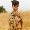 A young child in a field wears Sarah's Silks Animal Print Playsilk - Cheetah as a cape, with whiskers and a nose painted on their face, resembling a cheetah amid tall golden grass.