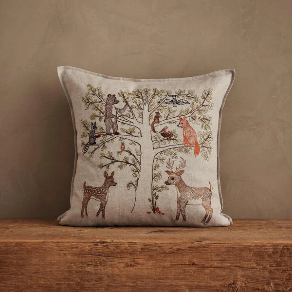 The Coral & Tusk Woodland Living Tree Pillow features embroidered deer, squirrel, fox, and birds around a family tree. Displayed against a beige wall on a wooden surface. Ships in one week.